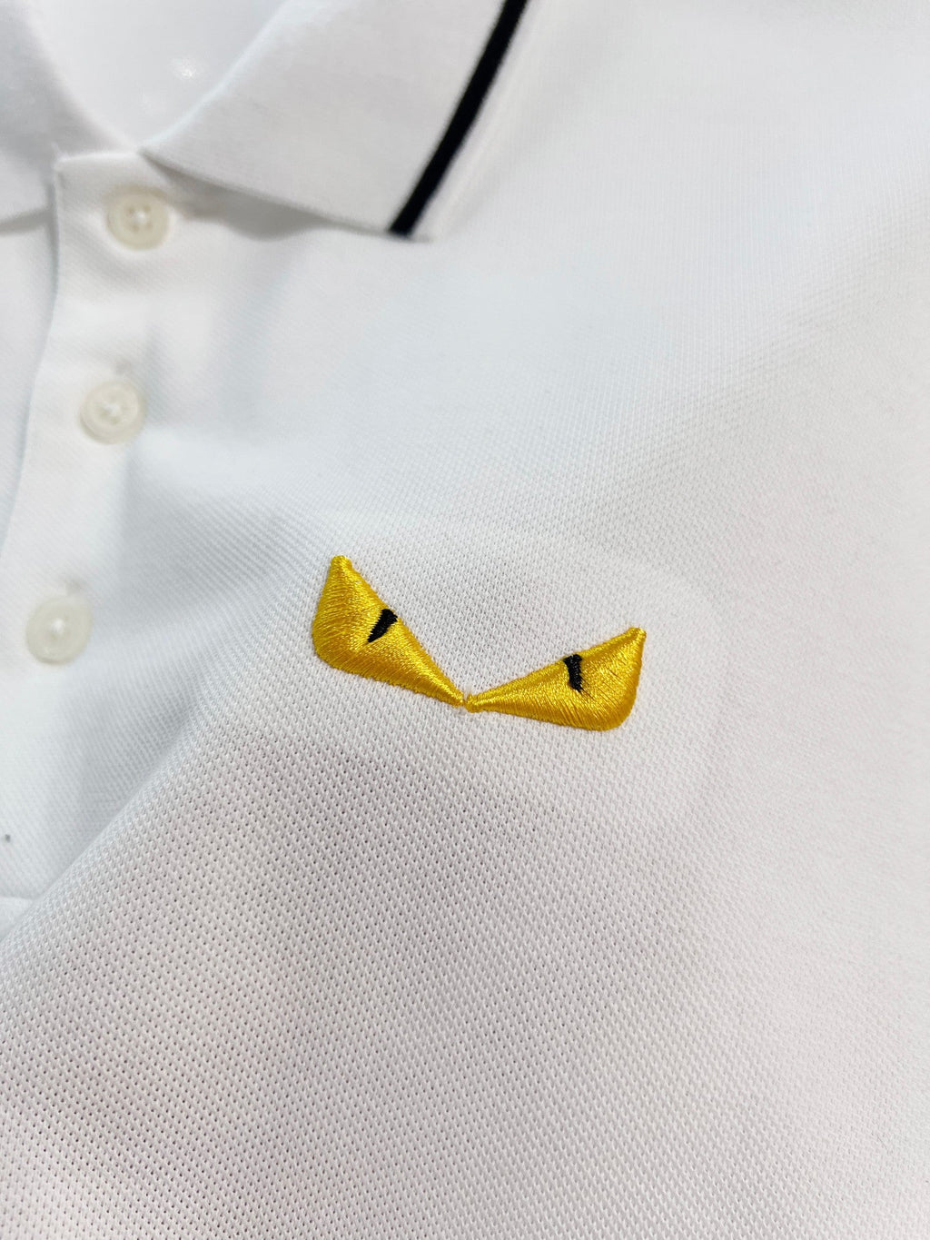 A polo shirt with embroidered letters on the eyes of a golden little monster on the chest